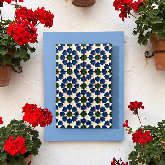 Vista general de frente del cuadro en relieve Patio de la Sultana de seis azulejos cerámicos en verde, azul cobalto y blanco montados sobre madera, colgado en pared de patio andaluz con flores rojas.